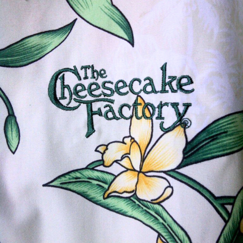 The Cheesecake Factory logo custom embroidered in deep green thread on patterned cream uniform fabric — professional restaurant embroidery by Embroidery Station LA, North Hollywood CA